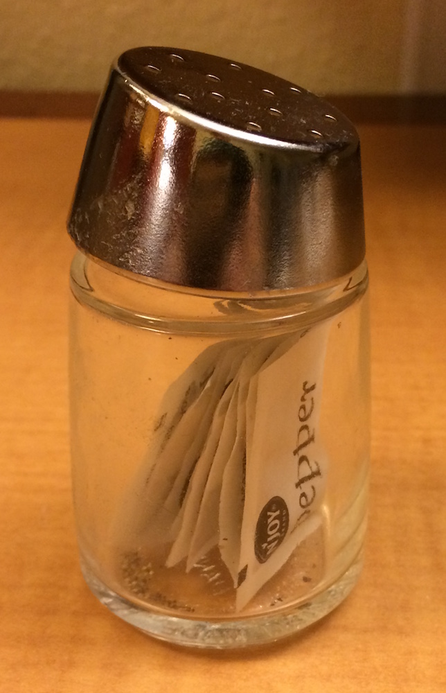 A photo of a glass pepper shaker. Instead of the pepper shaker containing pepper, it contains many packets of pepper.